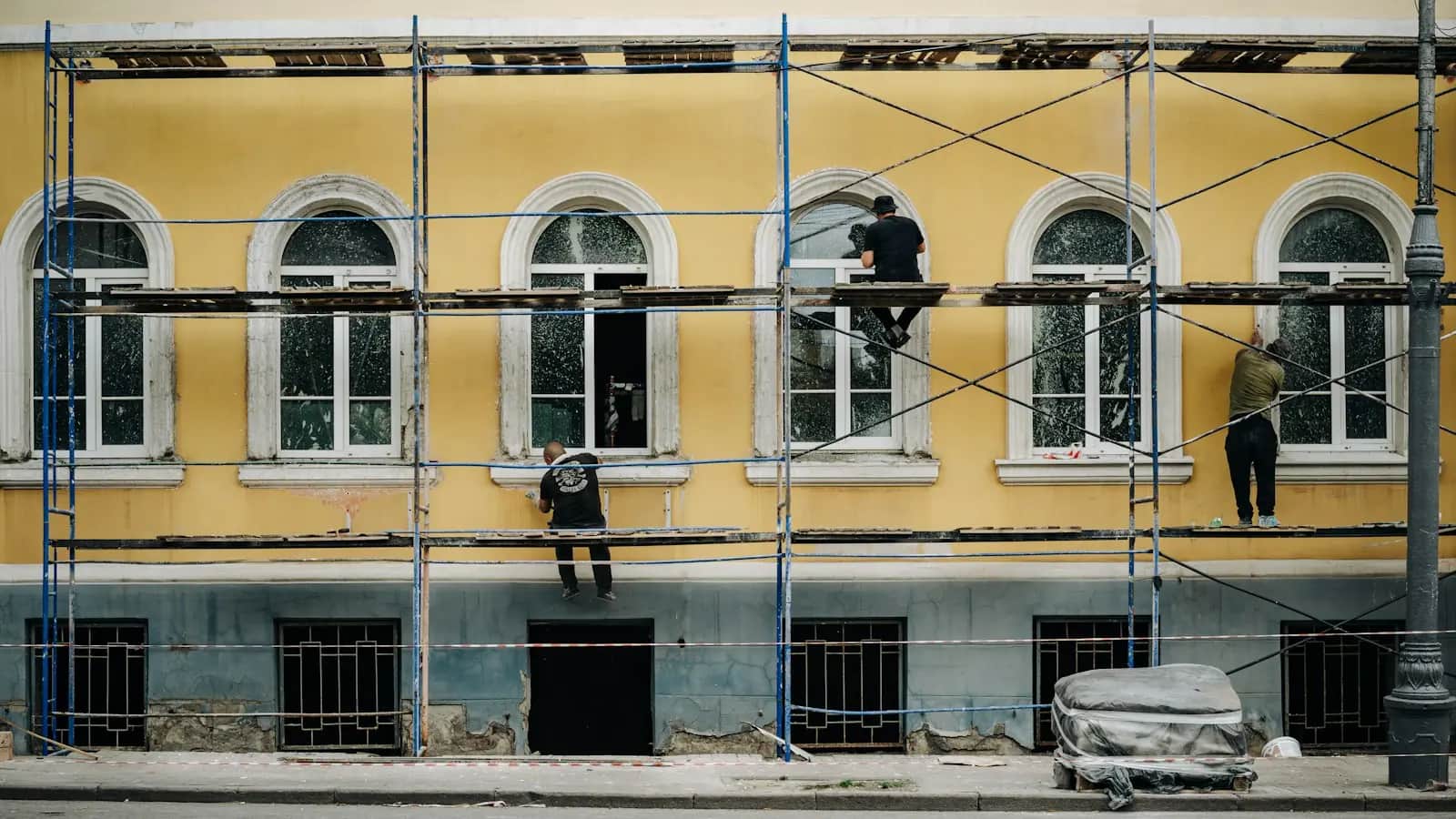 İskele üzerinde cephe onarımı yapan ustalar, sarı boyalı bir apartman cephesi